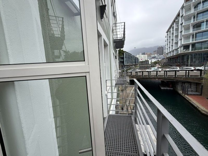 A narrow metal balcony overlooking a canal and bridge, featuring modern glass windows and industrial-style grated flooring.
