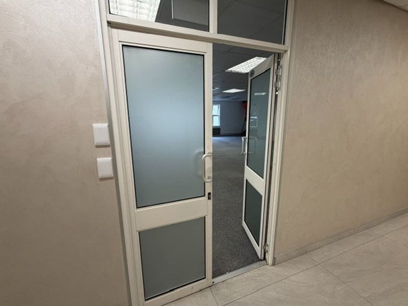 An open-plan office space viewed through frosted glass double doors, featuring neutral wall finishes, carpeted flooring, and a suspended ceiling with recessed lighting.