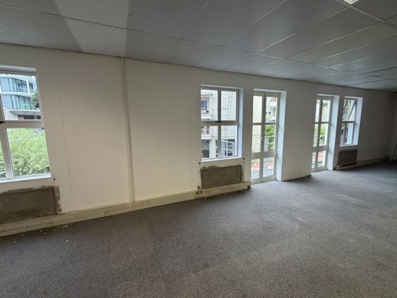 An empty office space featuring a suspended ceiling, carpeted flooring, and multiple windows providing natural light, currently requiring minor wall repairs.