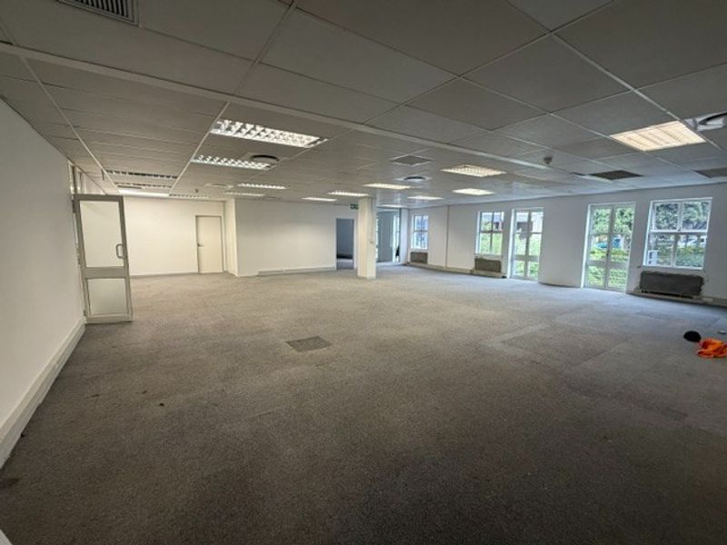 A spacious, open-plan office area featuring a suspended ceiling with recessed lighting, large windows providing natural light, and carpeted flooring.