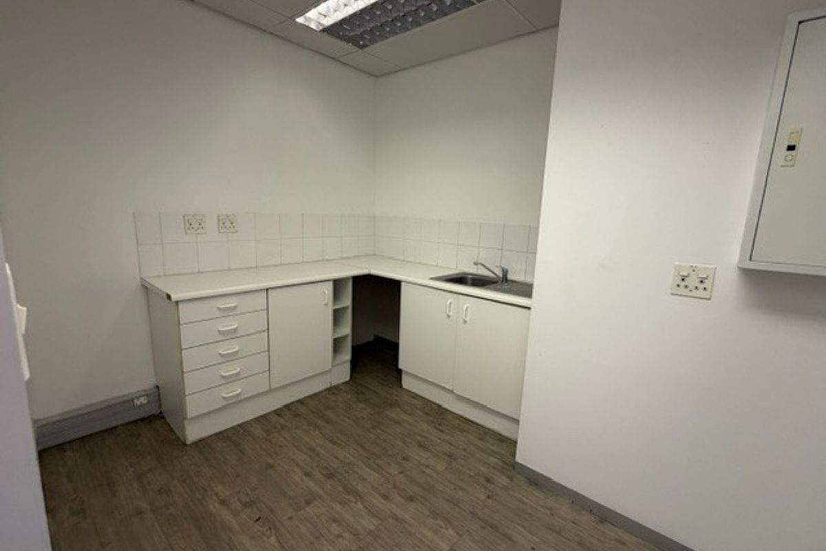 A functional office kitchenette featuring built-in white cabinetry, a stainless steel sink, tiled backsplash, and durable laminate flooring.