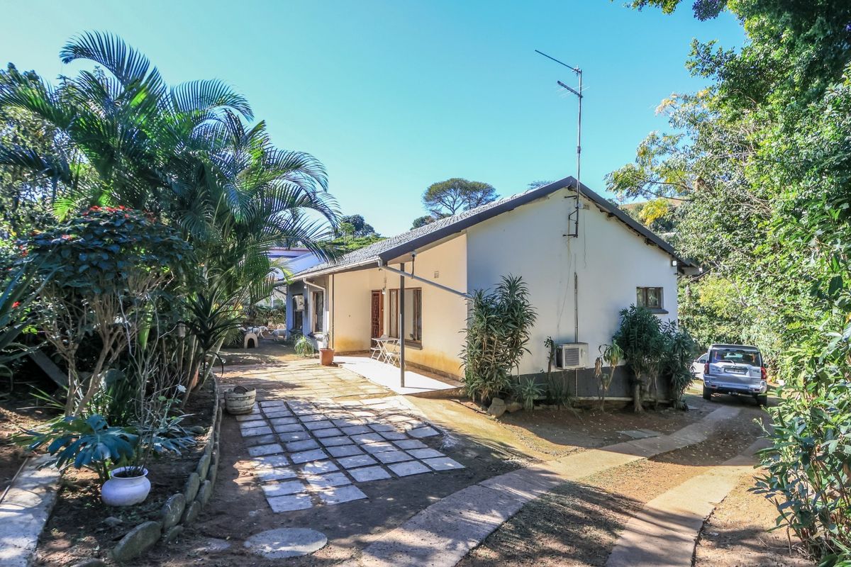 Rear view of the property featuring a paved walkway, established garden surroundings, and a concrete patio area adjacent to the main building structure.