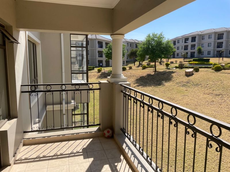 A covered balcony featuring tiled flooring and a decorative wrought iron balustrade, overlooking the complex's landscaped garden area.