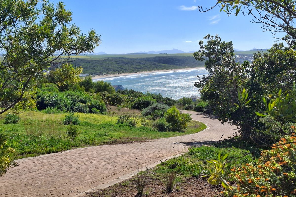 A scenic view of the coastline featuring a winding paved walkway through landscaped grounds, overlooking the ocean and distant hills.