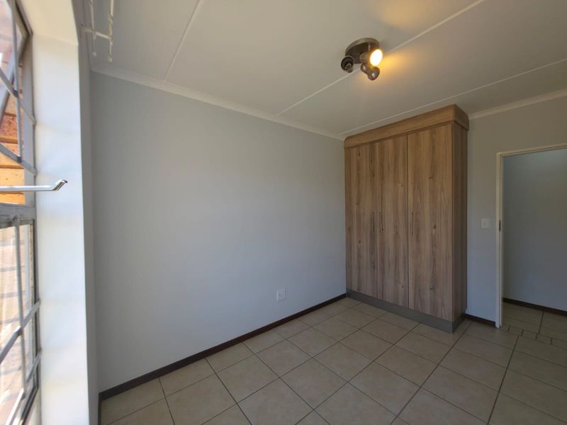 A clean, empty bedroom featuring neutral tiled flooring, a built-in wooden wardrobe, and a window with security bars.