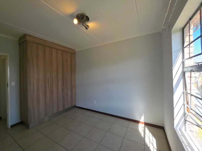 A bright, empty bedroom featuring neutral tiled flooring, built-in wooden cupboards, and a large window allowing for natural light.