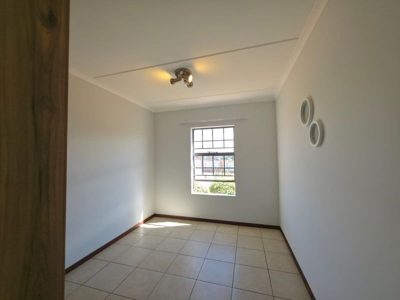 A bright, empty bedroom featuring neutral tiled flooring, a window with security bars, and a modern ceiling-mounted spotlight fixture.