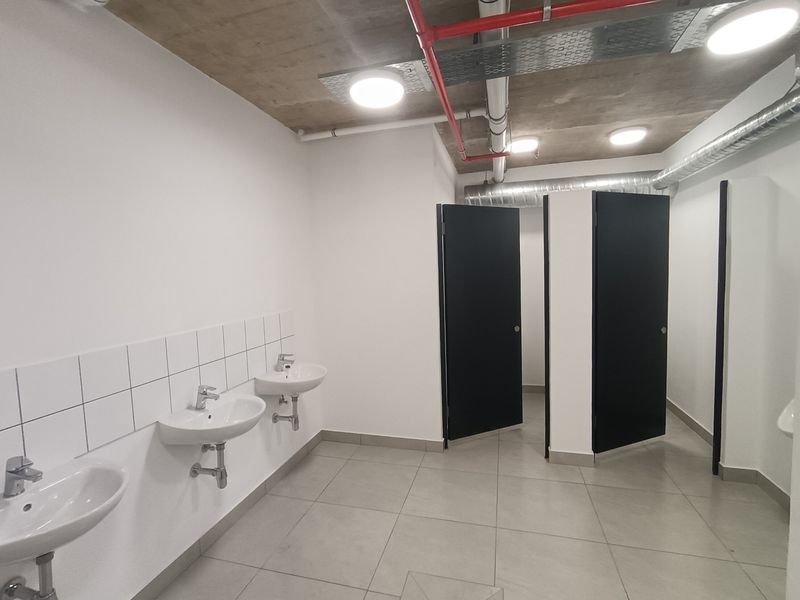 Modern industrial-style communal bathroom featuring multiple wall-mounted basins, tiled flooring, and exposed ceiling infrastructure with black cubicle doors.