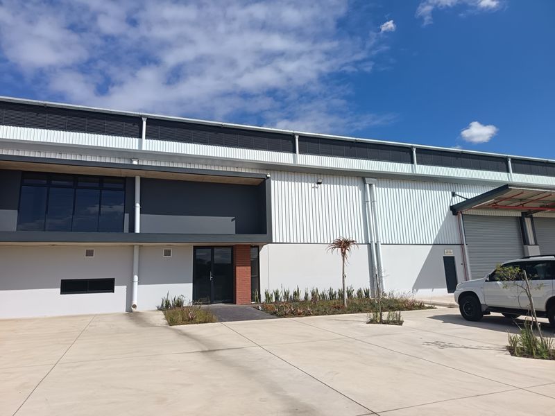 Modern industrial warehouse exterior featuring corrugated metal cladding, a large roller shutter door, and a paved concrete driveway with landscaped garden beds.