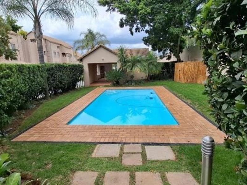 A well-maintained outdoor pool area featuring a clear swimming pool surrounded by brick paving and established garden landscaping.