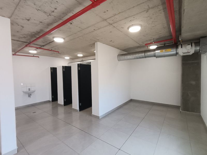 A modern, industrial-style bathroom facility featuring exposed concrete ceilings, red fire sprinkler piping, tiled flooring, and multiple private cubicles.