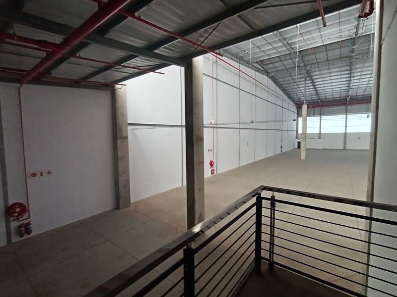 Spacious industrial warehouse interior featuring high ceilings, exposed fire suppression systems, concrete flooring, and structural columns with a mezzanine viewing area.