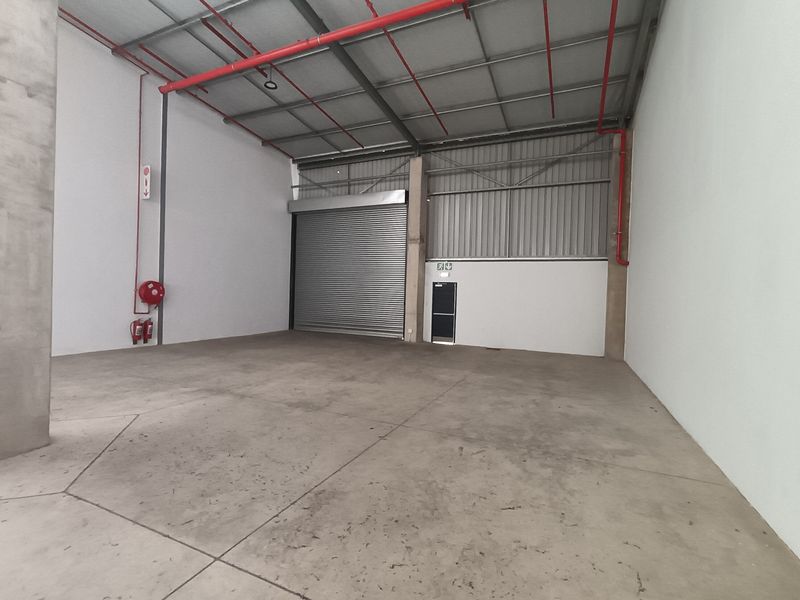 Spacious industrial warehouse unit featuring high ceilings, exposed fire suppression systems, concrete flooring, and a large roller shutter door for easy access.