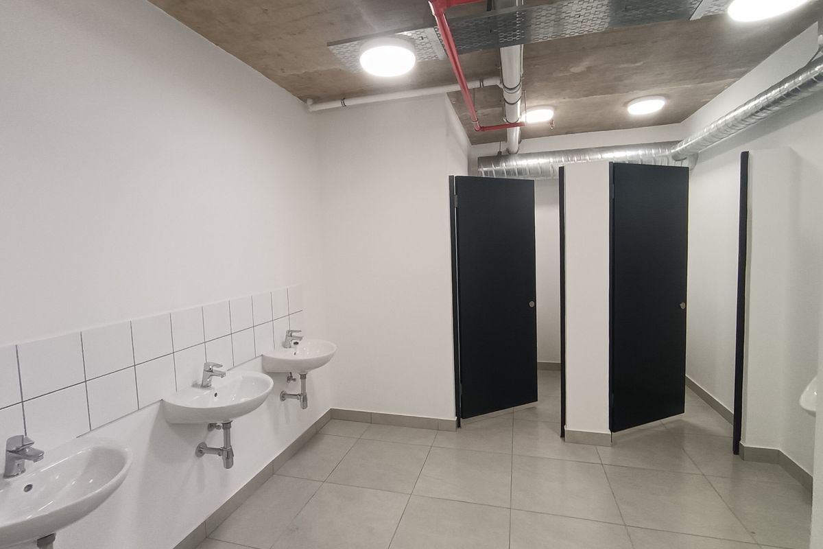 Modern industrial-style communal bathroom featuring multiple wall-mounted basins, tiled flooring, and exposed ceiling infrastructure with black cubicle doors.