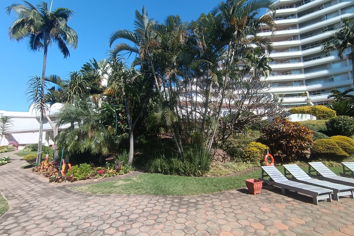 A well-maintained landscaped garden area with paved walkways and mature palm trees, situated in front of a multi-story apartment complex.