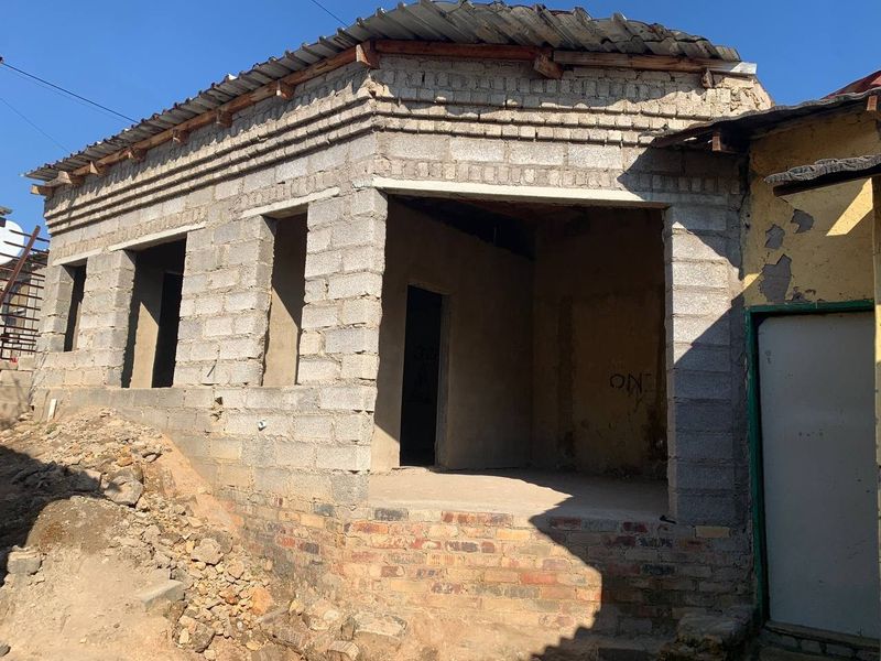 An unfinished residential structure featuring exposed concrete block walls, a corrugated metal roof, and a brick foundation, currently in a shell construction state.