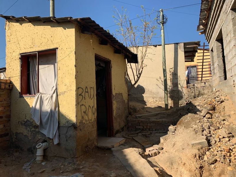 Exterior view of a small residential structure showing significant wear to the plaster walls and surrounding unlandscaped ground area.