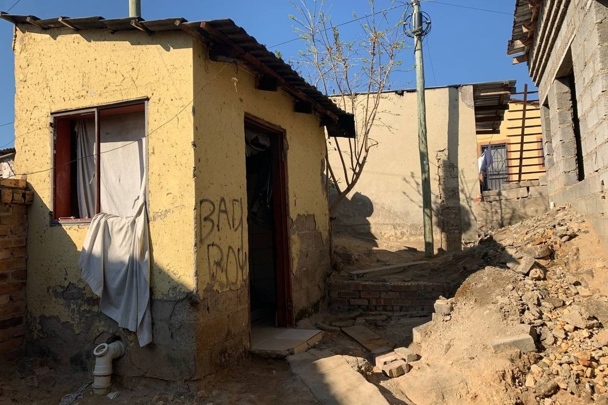 Exterior view of a small residential structure showing significant wear to the plaster walls and surrounding unlandscaped ground area.