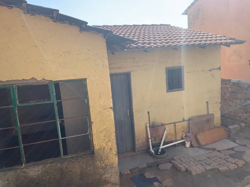 Rear exterior view of a house showing weathered yellow plaster walls, tiled roofing, and exposed external plumbing fixtures.