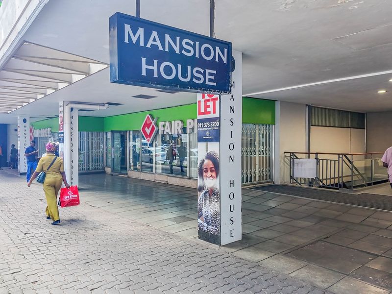 Front exterior view of Mansion House commercial building featuring a covered arcade, glass storefronts, and prominent signage along a paved pedestrian walkway.
