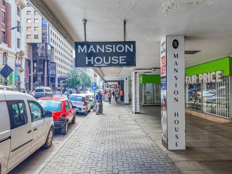 Street-level view of the Mansion House commercial building facade, featuring a covered pedestrian walkway, storefronts, and prominent signage in a busy urban setting.
