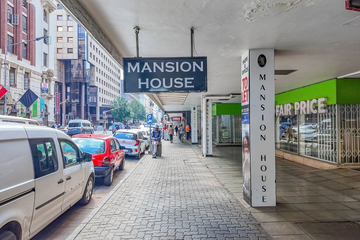 Street-level view of the Mansion House commercial building facade, featuring a covered pedestrian walkway, storefronts, and prominent signage in a busy urban setting.