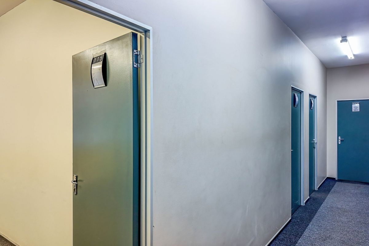 A commercial office hallway featuring multiple doors with signage, fluorescent lighting, and carpeted flooring.