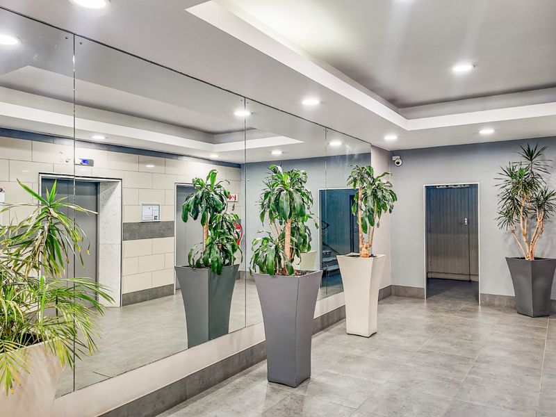 A modern commercial building lobby featuring tiled flooring, recessed ceiling lighting, a large mirrored wall, and elevator access.
