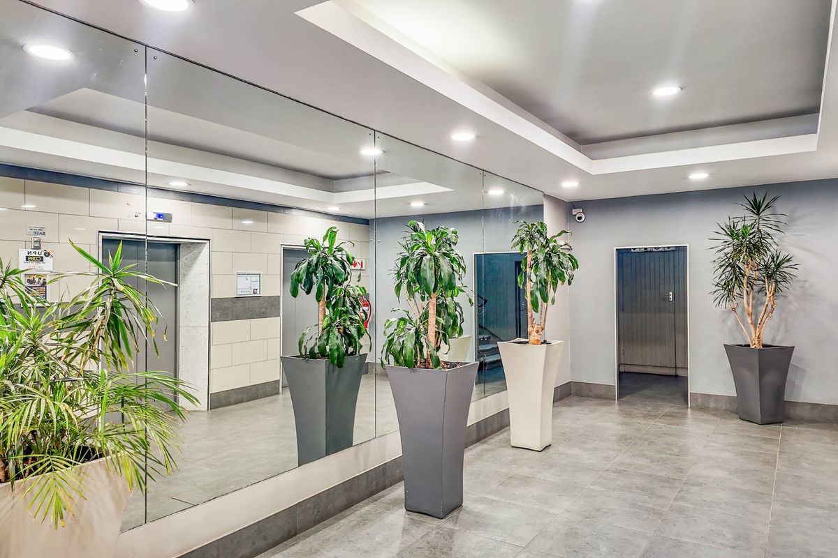 A modern commercial building lobby featuring tiled flooring, recessed ceiling lighting, a large mirrored wall, and elevator access.