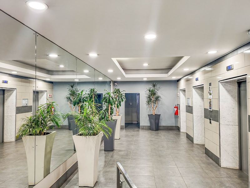 A modern commercial elevator lobby featuring tiled floors, recessed lighting, wall-to-wall mirrors, and multiple elevator access points.