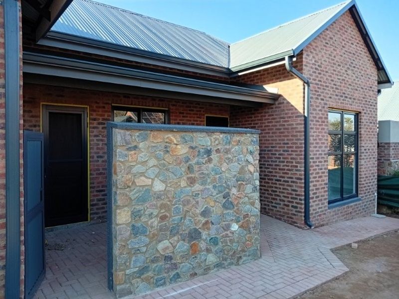 A well-maintained rear exterior view featuring a face brick facade, stone feature wall, corrugated metal roof, and paved patio area.