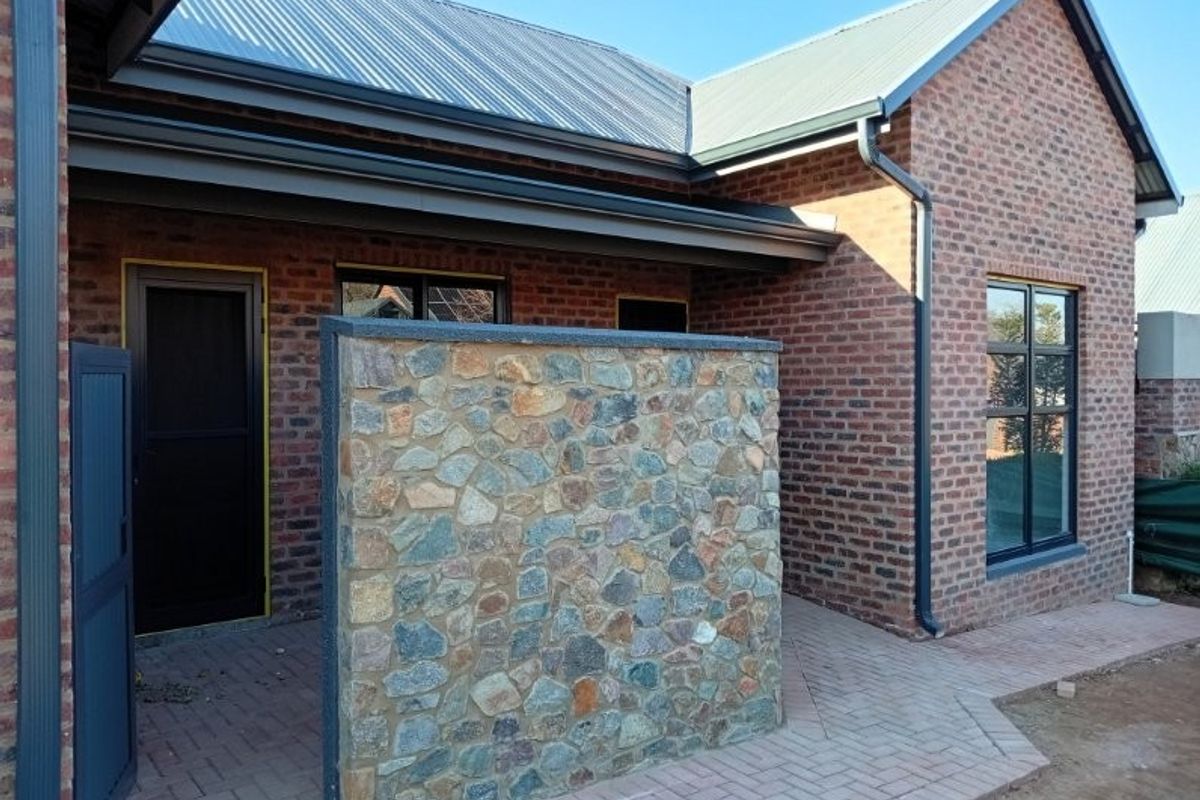 A well-maintained rear exterior view featuring a face brick facade, stone feature wall, corrugated metal roof, and paved patio area.