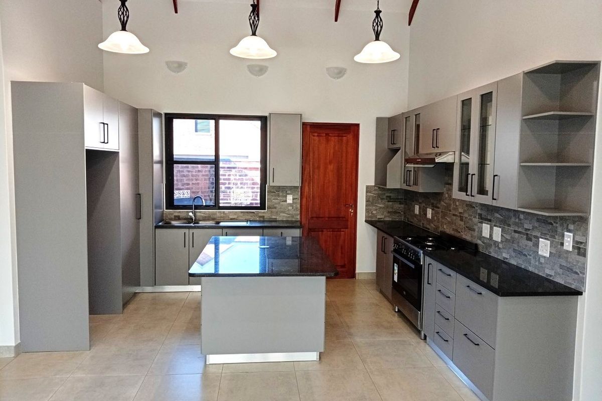 Modern kitchen featuring grey cabinetry, granite countertops, a central island, stone backsplash, and high ceilings with exposed rafters.