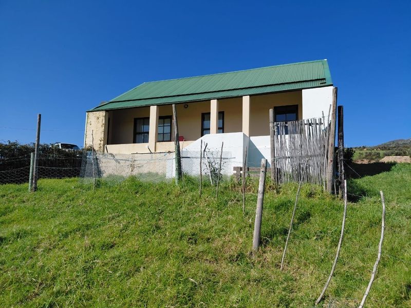 A single-story house featuring a green corrugated metal roof and a covered front veranda, set on a grassy slope with wire fencing.