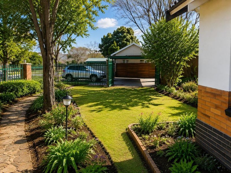 A well-maintained front garden featuring a stone pathway, manicured lawn, established garden beds, and secure perimeter fencing with a carport.