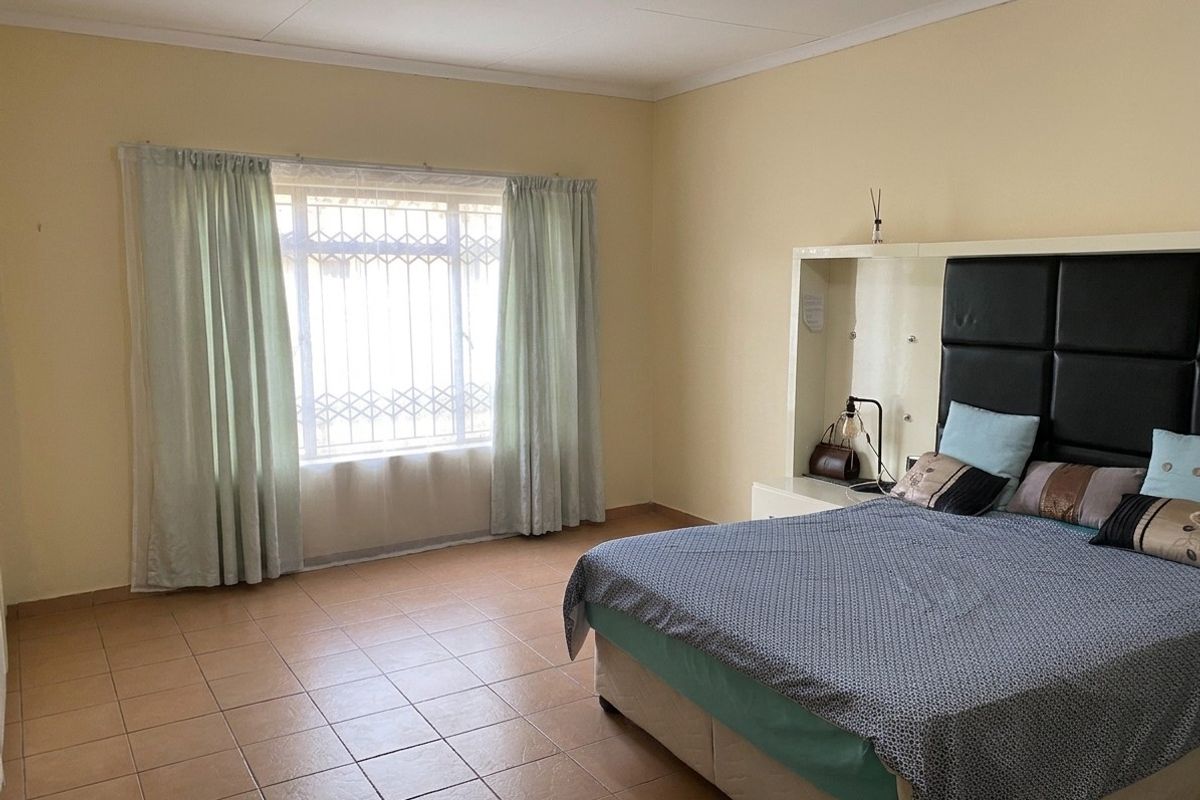 A spacious bedroom featuring tiled flooring, a built-in headboard unit, and a window fitted with a security gate.