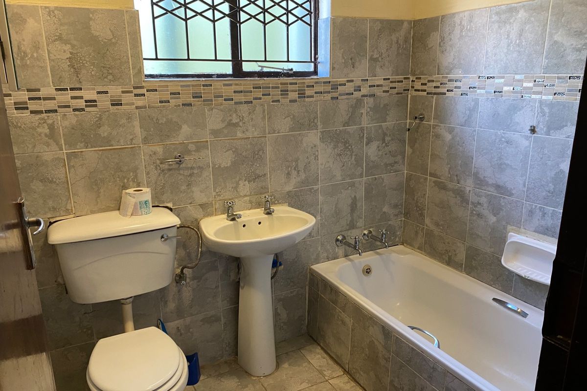 A functional bathroom featuring a pedestal basin, toilet, and bathtub with full-height wall tiling and a window with security bars.
