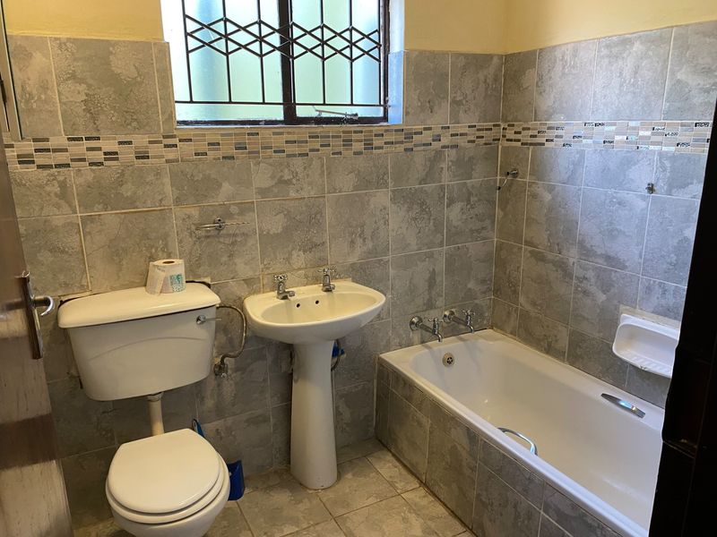A functional bathroom featuring a pedestal basin, toilet, and bathtub with full-height wall tiling and a window with security bars.