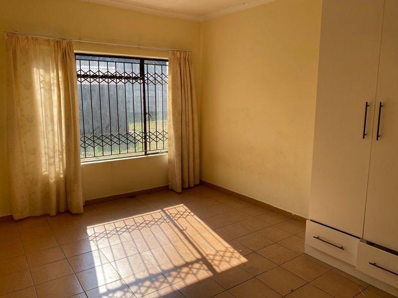 A bright, empty bedroom featuring tiled flooring, a built-in wardrobe, and a window fitted with a security gate.