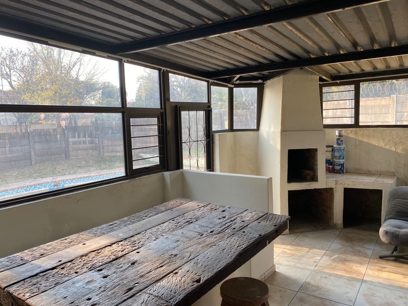Covered patio area featuring a built-in braai, tiled flooring, and large windows overlooking the garden and pool area.