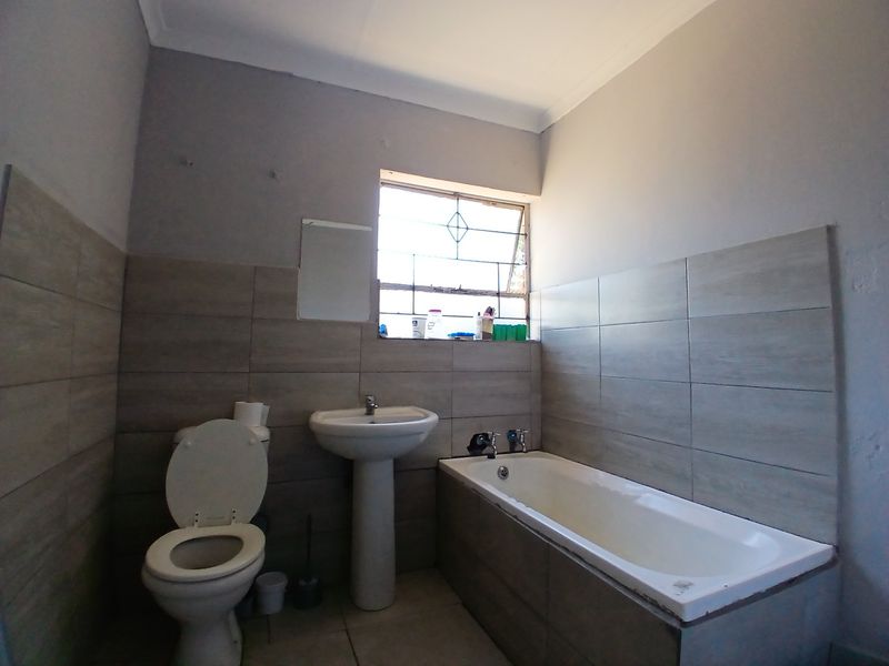 A functional bathroom featuring a pedestal basin, built-in bathtub, and toilet with neutral wall tiling and a window with security bars.