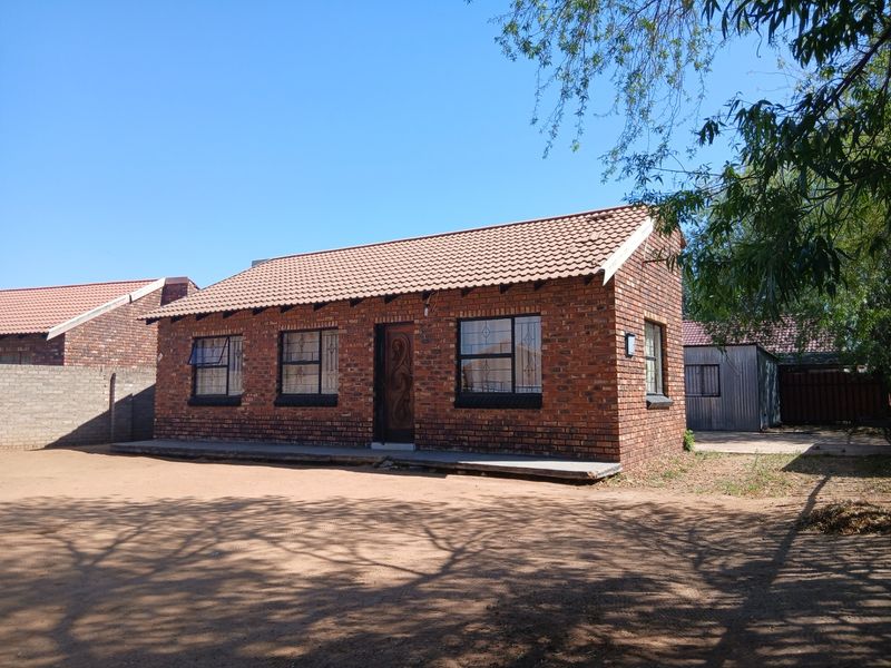A single-story face brick house featuring a tiled roof and security bars on windows, situated on a large, unpaved plot.