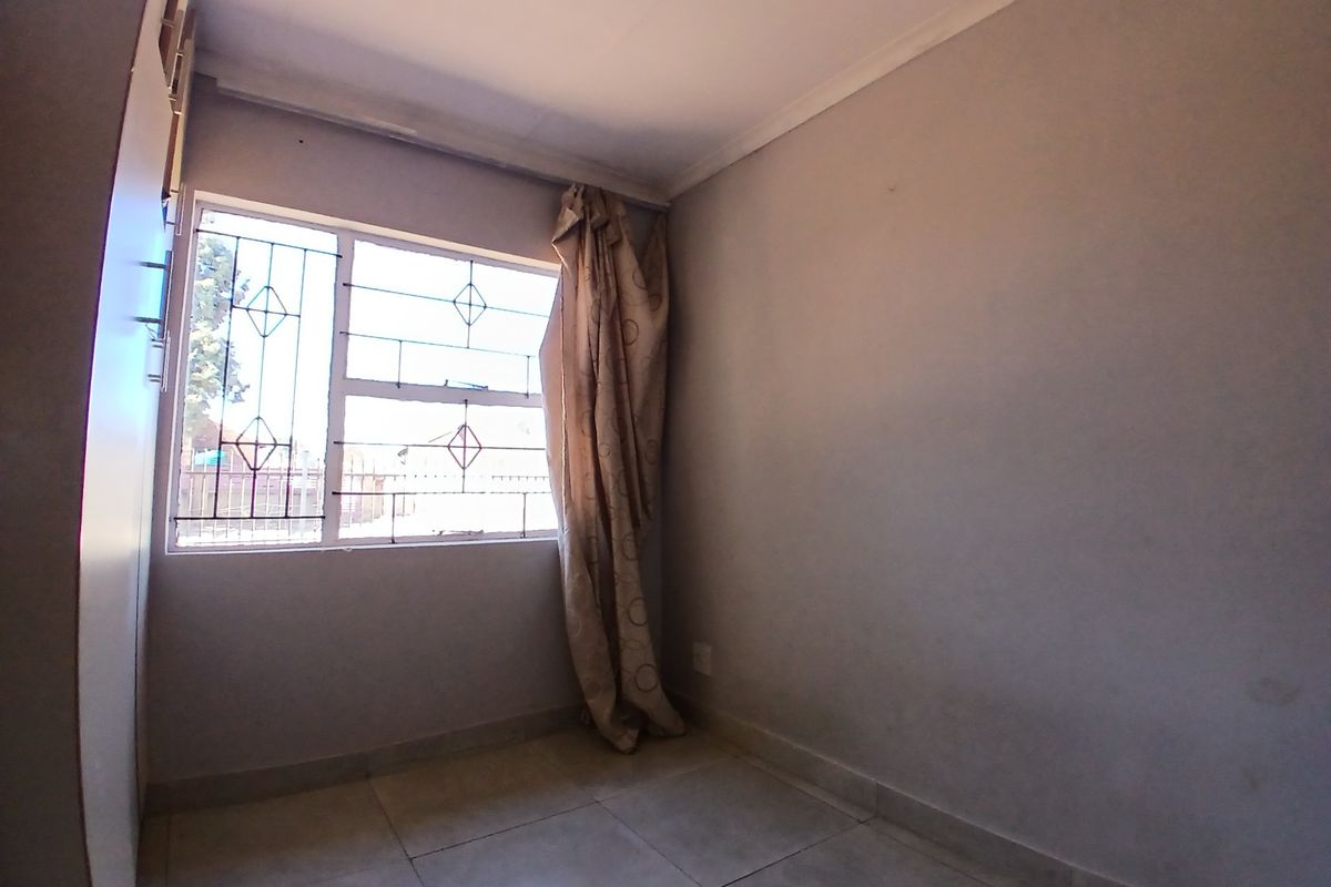 A compact, empty bedroom featuring tiled flooring, a built-in cupboard, and a window fitted with security bars.