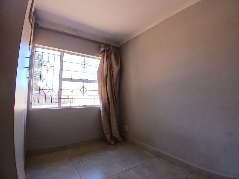 A compact, empty bedroom featuring tiled flooring, a built-in cupboard, and a window fitted with security bars.