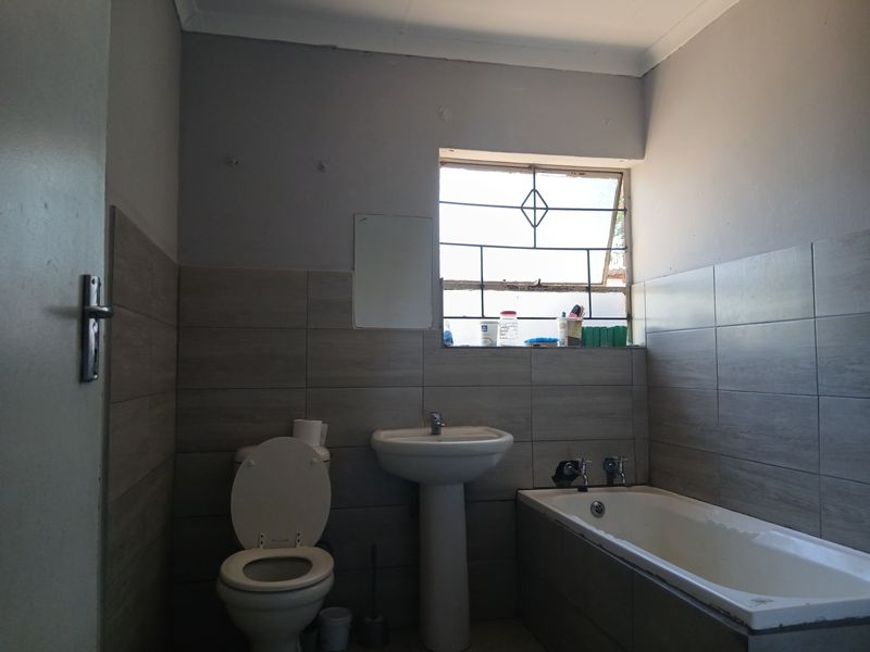 A functional bathroom featuring a pedestal basin, built-in bathtub, and toilet with neutral wall tiling and security bars on the window.