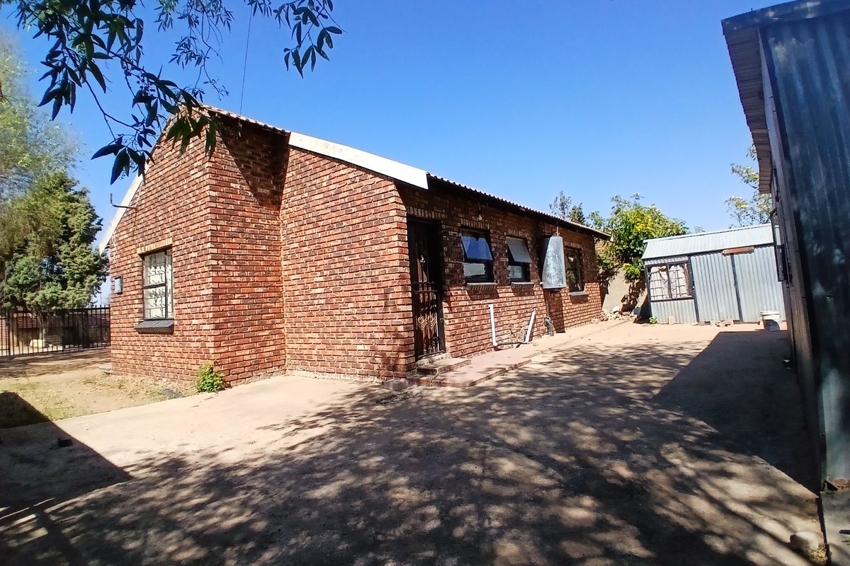 Rear view of a face brick house featuring a corrugated metal roof, security gate, and an adjacent metal outbuilding on a concrete driveway.