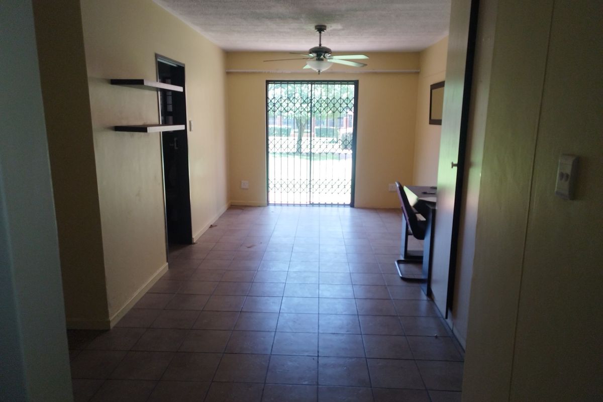 A sparsely furnished living area featuring tiled flooring, a ceiling fan, and a sliding glass door with a security gate leading outdoors.
