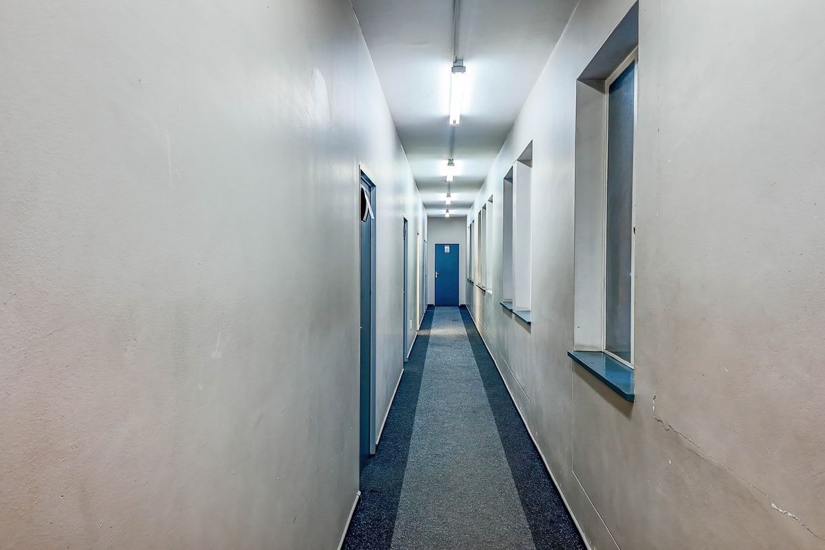 A long, functional commercial hallway featuring fluorescent strip lighting, carpeted flooring, and multiple access doors along the corridor.