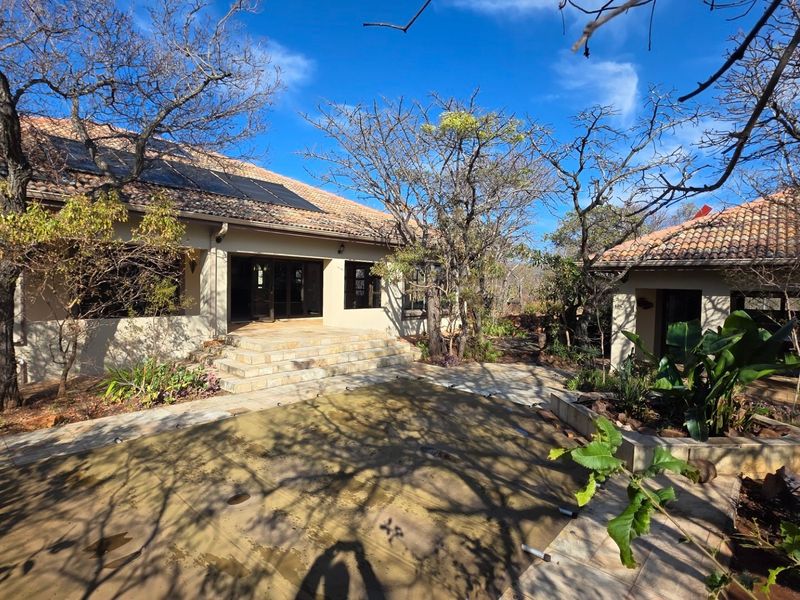 Rear exterior view featuring a stone patio, swimming pool with cover, and a well-maintained garden area with solar panels installed on the tiled roof.