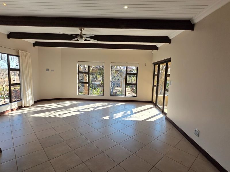 Spacious, light-filled living area featuring tiled flooring, exposed timber ceiling beams, and large windows with views of the garden.